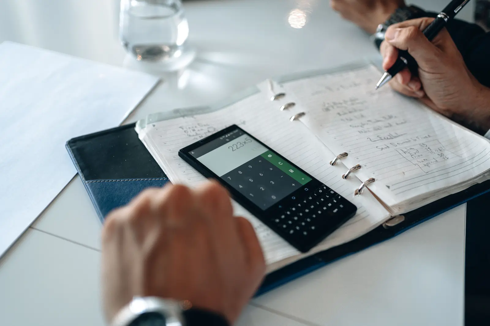 Hands writing in a notebook beside a phone calculator