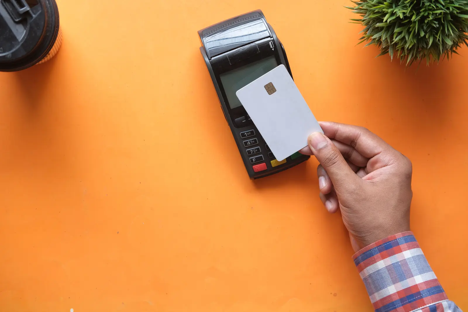 A hand holding a payment card over a card reader on an orange desk